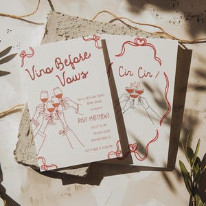 May include: Two white cards with red text and illustrations. One card reads "Vino Before Vows" and the other "Cir Cir!". Both cards feature line drawings of hands toasting wine glasses. The cards are for a bridal shower.
