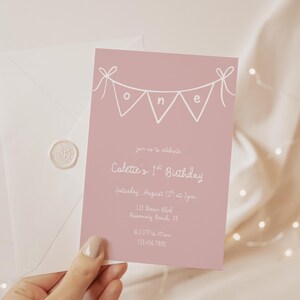 May include: A pink birthday invitation with white text and a banner spelling "one". The invitation reads "Colette's 1st Birthday" and includes event details. An envelope with a wax seal is in the background.