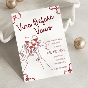 May include: A white invitation card with burgundy text and illustrations. The text reads "Vino Before Vows" and includes details for a wine tasting bridal shower. The illustration shows hands toasting with wine glasses.