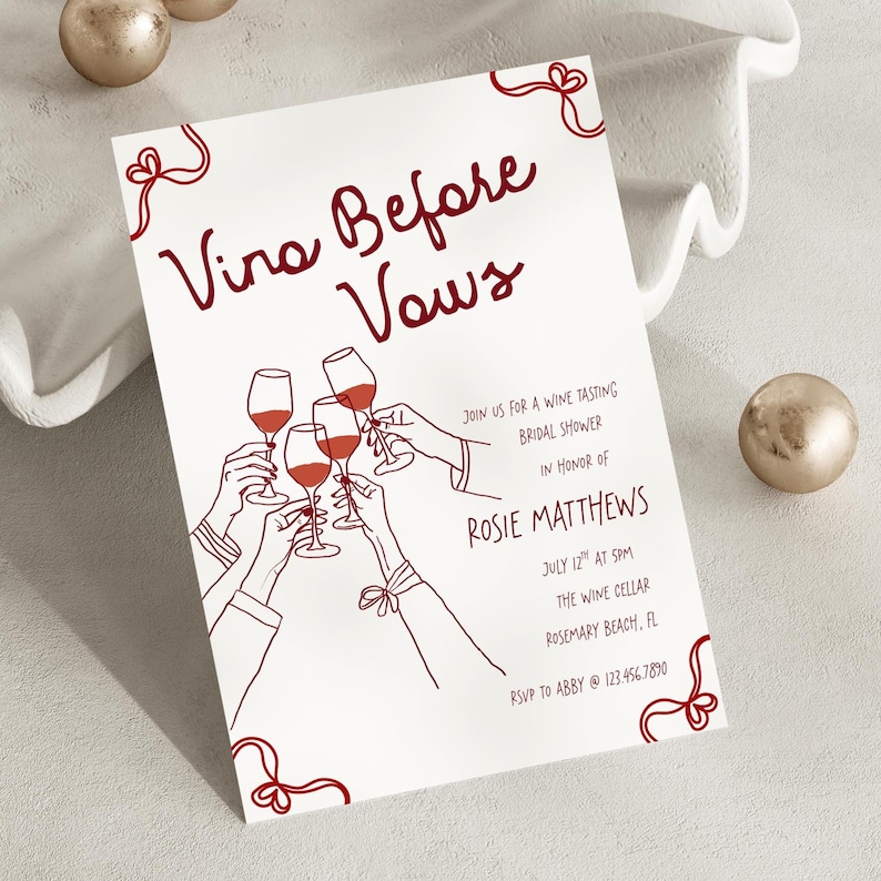 May include: A white invitation card with burgundy text and illustrations. The text reads "Vino Before Vows" and includes details for a wine tasting bridal shower. The illustration shows hands toasting with wine glasses.
