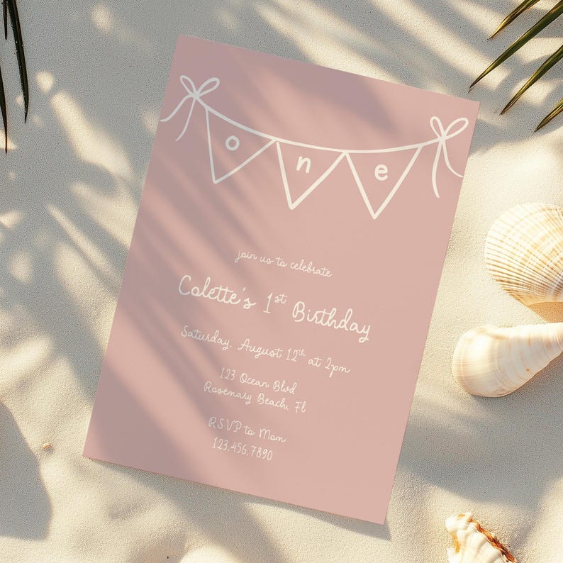 May include: A pink birthday invitation card with white text and a banner reading "one". The card is for Colette's 1st birthday, including date, time, and location. The card rests on a sandy surface.