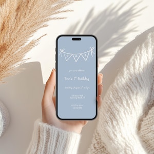 May include: A smartphone screen shows a light blue birthday invitation. The invitation reads "Finn's 1st Birthday" with event details. The phone is held by a person wearing a white sweater. Dried plants are in the background.