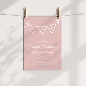 May include: A pink birthday invitation card with white text and a banner spelling "ONE". The invitation reads "Colette's 1st Birthday" and includes the date, time, and location. The card is held by two wooden pegs.
