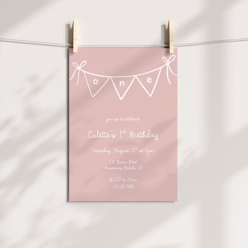 May include: A pink birthday invitation card with white text and a banner spelling "ONE". The invitation reads "Colette's 1st Birthday" and includes the date, time, and location. The card is held by two wooden pegs.