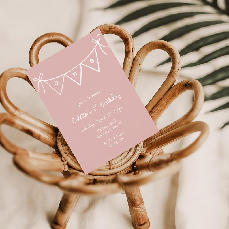 May include: A pink birthday invitation with white text and a banner design that reads "ONE". The invitation is for Colette's 1st Birthday, with the date, time, and location details. The invitation is resting on a woven chair.
