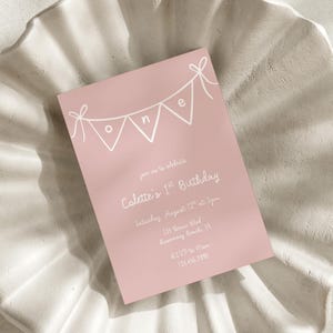 May include: A pink birthday invitation with white text and a banner spelling "ONE". The invitation reads "Join us to celebrate Colette's 1st Birthday" with the date, time, and location. RSVP details are also included.