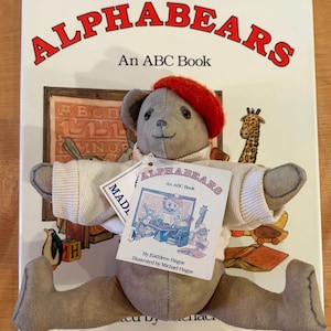 May include: A gray plush teddy bear wearing a red beret and a cream-colored ribbed sweater, positioned over the book Alphabears: An ABC Book by Kathleen Hague, illustrated by Michael Hague, which features a colorful illustration of animals.