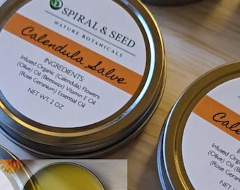 May include: Round metal tins of Spiral & Seed Calendula Salve with white labels featuring orange accents. The labels list ingredients including organic calendula flowers, olive oil, beeswax, vitamin E oil, and rose geranium essential oil, net wt. 2 oz.