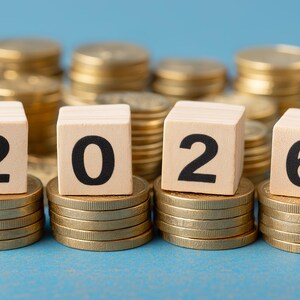 May include: Stacks of gold coins and wooden blocks spelling out the year 2026. The blocks sit atop smaller coin stacks, with additional coin stacks in the background against a blue backdrop. Financial concept.