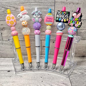 May include: A collection of Easter-themed pens in a clear acrylic holder. Each pen features a unique design, including Easter eggs, bunnies, and the words "Happy Easter." The pens have colorful barrels in yellow, orange, white, blue, pink, and purple.