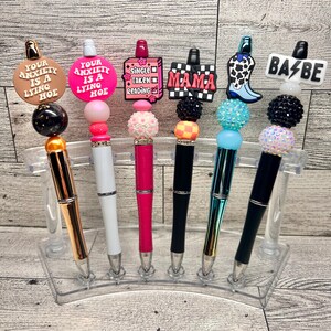 May include: A collection of six decorative pens with various toppers. The toppers include phrases like "Your Anxiety is a Lying Hoe," and "Mama," along with other designs. The pens are displayed in a clear plastic holder.
