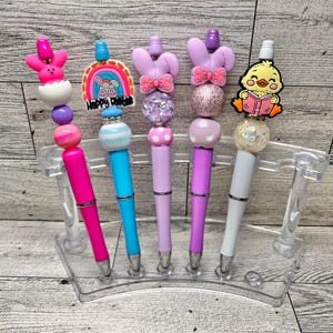 May include: A set of five decorative pens in a clear plastic holder. Each pen features a different Easter-themed topper, including a bunny, a rainbow, and a reading chick. The pens have colorful barrels in pink, blue, and purple.