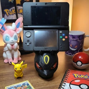 Puede incluir: Una consola de juegos portátil Nintendo 3DS negra se muestra sobre un soporte negro con temática Umbreon. También se ven una figura de Pikachu, un peluche de Sylveon, una Poké Ball y un cuaderno con temática Pokémon. Una taza con un diseño de Gengar está en el fondo.