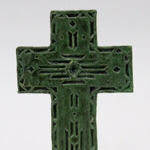 May include: A green cross with a detailed geometric pattern. The cross has a textured surface and a dark green color. The design features diamond and rectangular shapes. The cross is a religious symbol.