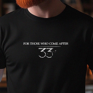 May include: Black t-shirt with the text "FOR THOSE WHO COME AFTER 33" in white. The shirt is short-sleeved and appears to be made of a soft material. The image shows the shirt on a person.