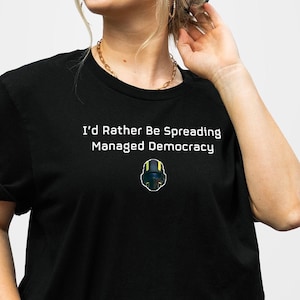Puede incluir: Una camiseta negra con texto blanco que dice "I'd Rather Be Spreading Managed Democracy" y un pequeño gráfico de una persona con casco.