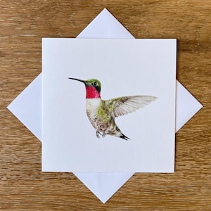May include: A square white greeting card with a watercolour painting of a hummingbird with outstretched wings. The bird has a red throat, green head, and speckled body. The card is inside a white envelope, set against a wooden background.