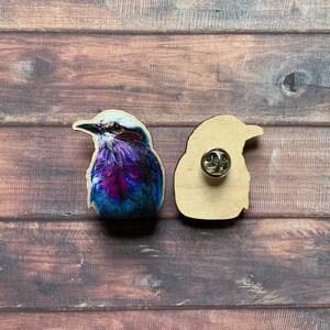 May include: A wooden pin featuring a colourful bird with blue, purple and white feathers. The back of the pin is plain wood with a metal pin back.