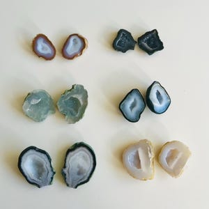 May include: A selection of polished geode slices, showcasing a variety of colours. The geodes feature shades of brown, blue, green, and white, with crystalline interiors. The natural stone formations are arranged on a white surface.