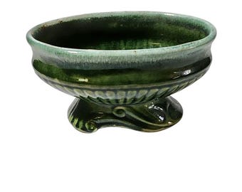 May include: A green ceramic planter with a scalloped rim and a decorative base. The planter is oval shaped and has a light green glaze.