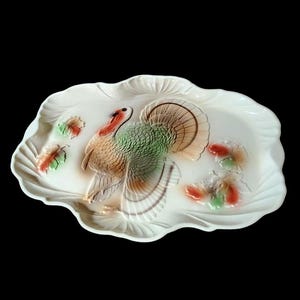 May include: A white ceramic serving platter with a raised design of a turkey with green, brown, and orange feathers. The turkey is surrounded by three green and red leaves.