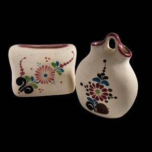 May include: Two hand-painted ceramic vases with floral designs. The larger vase has a handle and a round body. The smaller vase is rectangular and has a flat base.