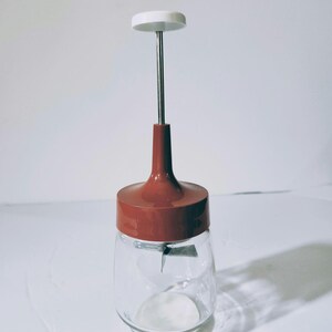 May include: A vintage food chopper with a clear glass jar, a dark red lid, and a white handle. The chopper has a stainless steel blade inside the jar and a long stainless steel rod connecting the handle to the lid.
