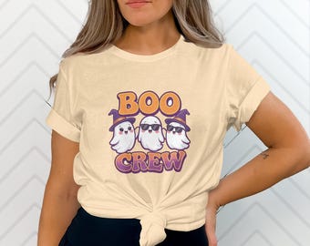 Può includere: T-shirt color crema con la scritta "BOO CREW" in arancione e viola, con tre fantasmi dei cartoni animati. Due fantasmi indossano cappelli da strega e uno indossa occhiali da sole. La maglietta è annodata in vita.