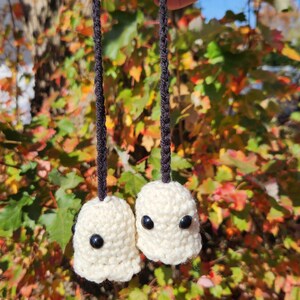 May include: Two small, off-white crocheted ghost figures with smooth black bead eyes. Each ghost is attached to a long, thin, black crocheted cord, allowing them to hang together. The figures feature a rounded, textured yarn construction.