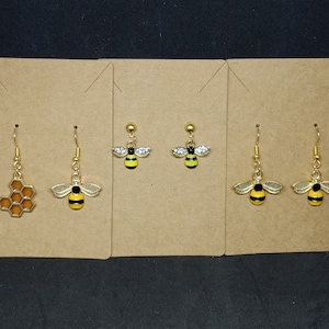 May include: A collection of gold-toned earrings featuring bee and honeycomb designs. The earrings include a honeycomb shape, bee pendants with yellow and black stripes, and bee pendants with rhinestone wings. Each earring has a gold-colored hook.