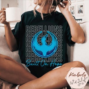 May include: Black t-shirt with the word "REBELLIONS" in a white outlined font. A blue phoenix graphic is centered on the shirt. The text "Built On Hope" is below the graphic. The person is holding a white mug.