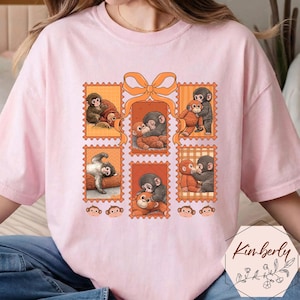 May include: Pale pink t-shirt with a design of postage stamps featuring illustrations of monkeys embracing. The stamps are orange with an orange bow. The shirt has a loose fit.