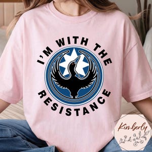 May include: A light pink t-shirt with the text "I'M WITH THE RESISTANCE" in black, surrounding a blue and white emblem featuring a black bird. The shirt is a casual wear item.