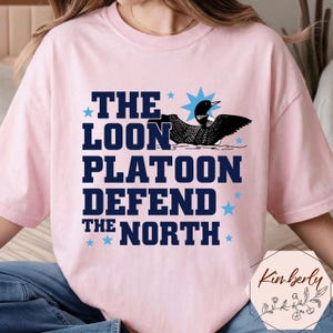 May include: A light pink t-shirt with the text "THE LOON PLATOON DEFEND THE NORTH" in dark blue. A black and white loon graphic is above the text. Blue stars are scattered around the text.