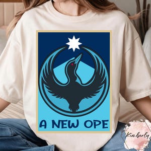 May include: Cream-colored t-shirt featuring a graphic design with a bird-like silhouette in shades of blue and a white star. The text "A NEW OPE" is printed in blue at the bottom of the design. The shirt is a casual wear item.