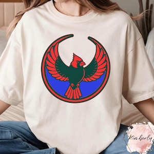 May include: Cream-colored t-shirt featuring a stylized cardinal design. The bird is depicted with red wings and head, a green body, and is set against a blue circle with red and green borders. The design is centered on the shirt.