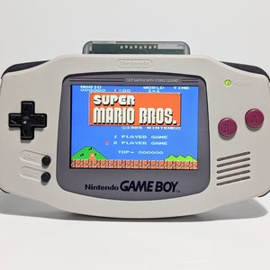 May include: A white and gray Game Boy Color handheld gaming console with a black d-pad and buttons. The screen displays the game Super Mario Bros. with the text "1985 Nintendo" and "1 Player Game" visible.