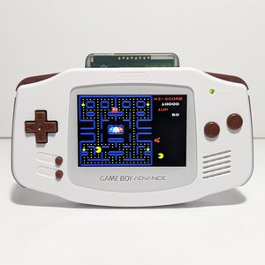 May include: A white Game Boy Advance handheld console with brown accents and a Pac-Man game displayed on the screen. The console has a cartridge inserted at the top and features buttons and a directional pad.