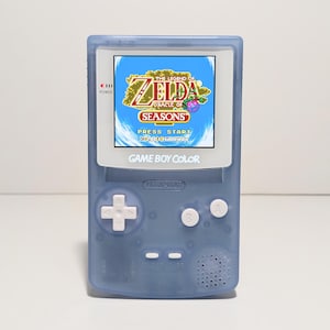 May include: A blue Game Boy Color handheld video game console with a white faceplate. The screen displays the game "The Legend of Zelda: Oracle of Seasons".