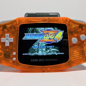 May include: An orange Game Boy Advance handheld gaming console with a clear plastic shell. The screen displays the title screen for the game "Mega Man Zero 4". The text "PRESS START" is visible on the screen.