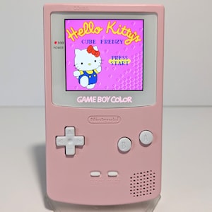 May include: A pink Game Boy Color handheld console displaying the "Hello Kitty's Cube Frenzy" game. The screen shows Hello Kitty in a blue outfit. The console has a directional pad, A and B buttons, and a power indicator.