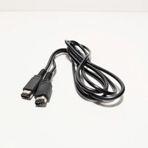 May include: Black video game controller cable with two connectors. The cable is coiled and lies on a white surface. The connectors are rectangular with multiple pins.