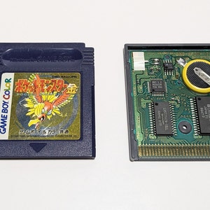 Op de afbeelding: Een close-up van een Game Boy Color-cartridge voor de Japanse versie van Pokemon Gold. De cartridge is blauw met een gouden label waarop een afbeelding van de legendarische Pokemon Ho-Oh staat. De achterkant van de cartridge toont de printplaat en de batterij.