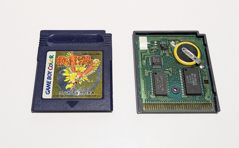Op de afbeelding: Een close-up van een Game Boy Color-cartridge voor de Japanse versie van Pokemon Gold. De cartridge is blauw met een gouden label waarop een afbeelding van de legendarische Pokemon Ho-Oh staat. De achterkant van de cartridge toont de printplaat en de batterij.