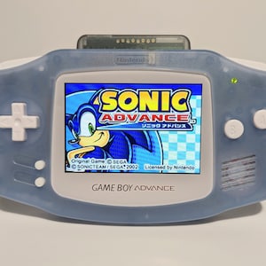 May include: A light blue Game Boy Advance handheld console with a game cartridge inserted. The screen displays the "Sonic Advance" game logo featuring Sonic the Hedgehog. The console has white buttons and a directional pad.