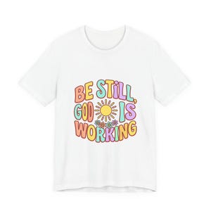 Puede incluir: Camiseta blanca con un gráfico colorido que dice "BE STILL, GOD IS WORKING". El diseño presenta un sol, flores y texto en tonos naranja, amarillo, azul y morado. Una camiseta casual para todos los días.