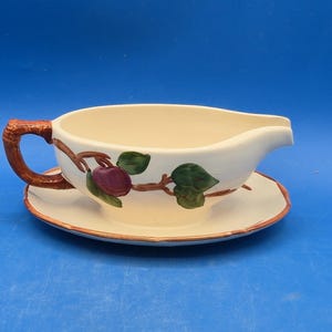 May include: A cream-colored ceramic gravy boat with an attached saucer, featuring a brown branch-shaped handle and a painted design of a purple plum with green leaves. The rim of the saucer and the handle have a brown painted trim detail.
