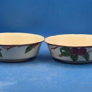 May include: Two round ceramic bowls with a cream-colored interior and a white exterior. The exterior features a hand-painted pattern of purple plums with green leaves and brown branches. The rims of the bowls are finished with a thin brown line.