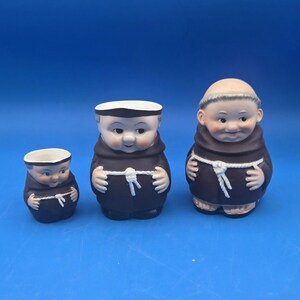 May include: Set of three ceramic monk figurines. The figurines are in different sizes and are all wearing brown robes with white rope belts. The largest figurine has a bald head, while the other two have white caps.