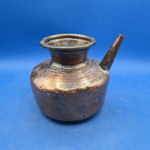 May include: A vintage copper vessel with a wide, circular opening and a single angled spout. The exterior features a weathered patina with dark oxidation spots and decorative etched patterns around the neck and upper body of the container.